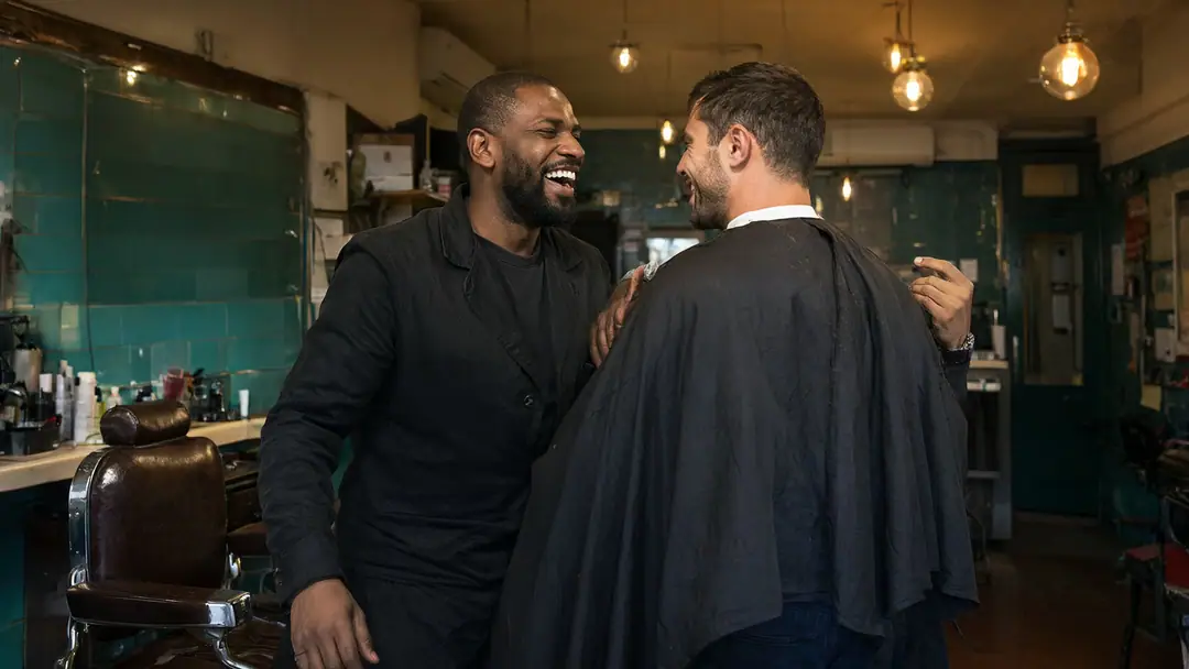 A barber and his customer sharing a laugh after a haircut in a warmly lit barbershop.