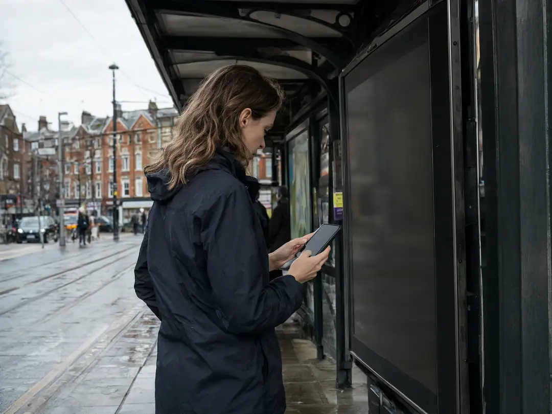 A woman checks her phone at a covered tram stop on a rainy day.