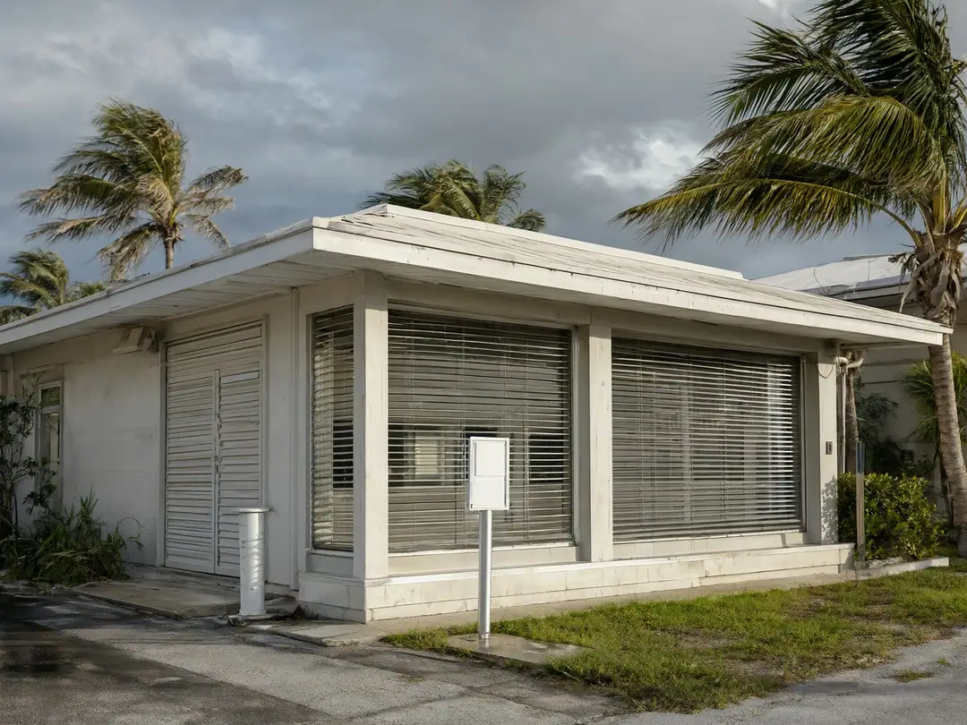 A small South Florida office building with hurricane shutters partly closed under a stormy sky.