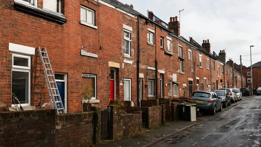A narrow street of red brick terraced houses on a cloudy day, with a ladder leaning against one front wall.