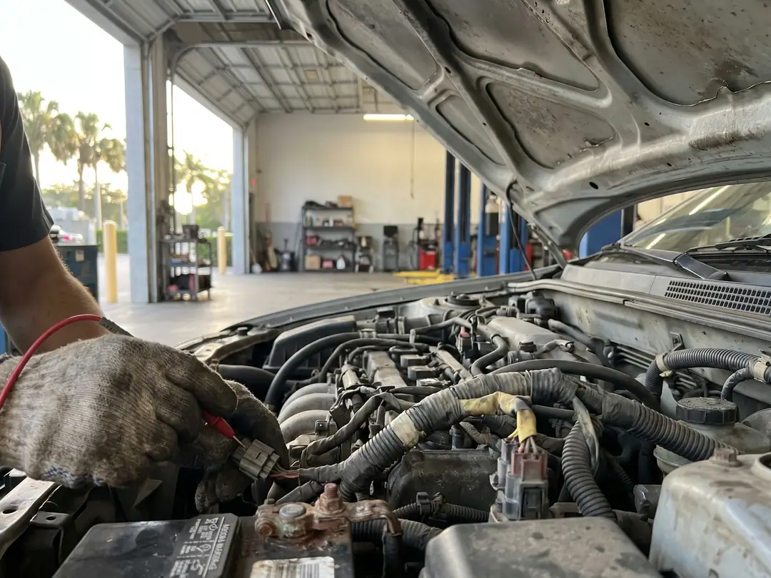 A gloved hand testing engine wiring with a multimeter under an open car hood in a service bay.