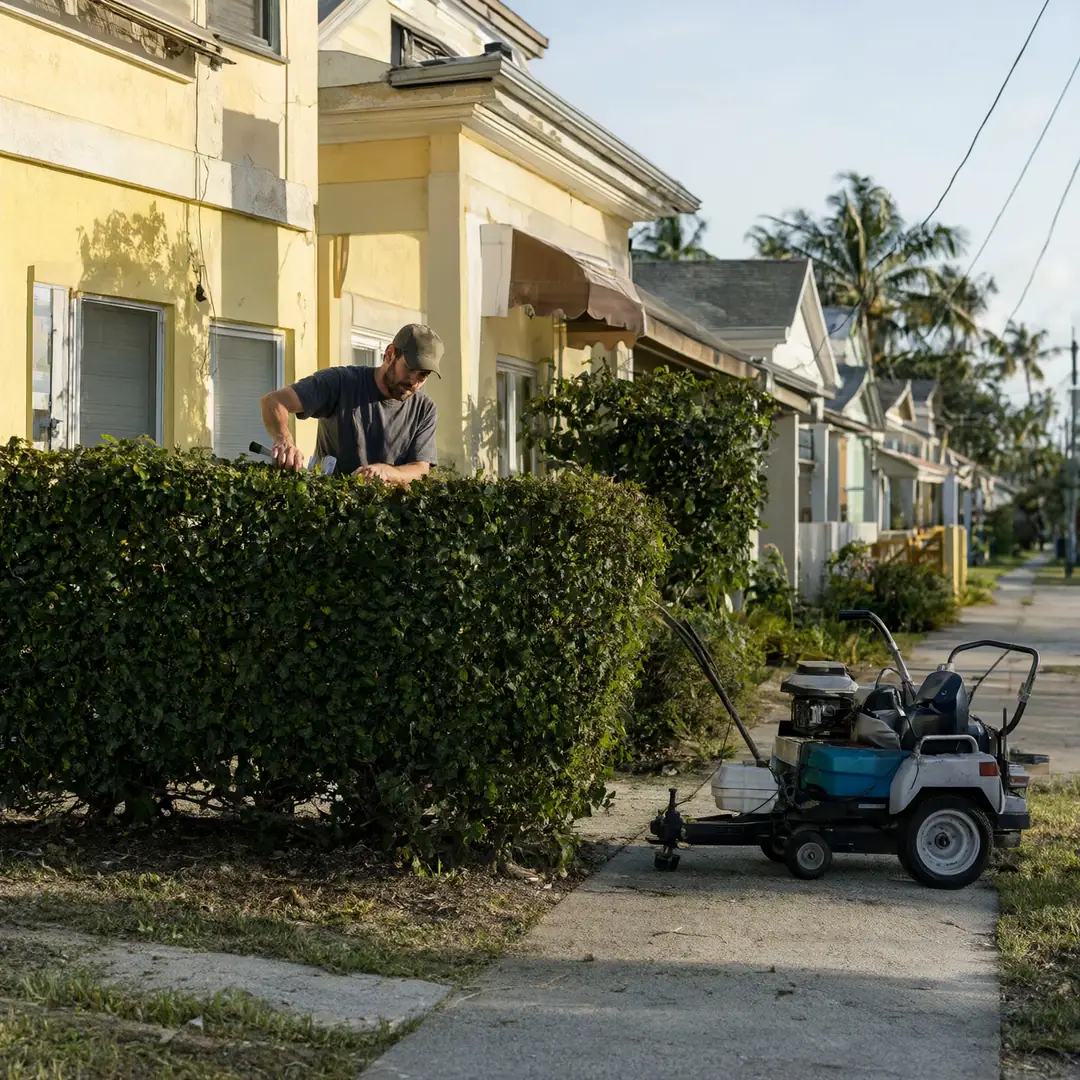 A landscaper trimming a hedge outside a yellow house with his mower parked by the sidewalk.